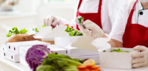 Twee medewerkers in een professionele keuken met rode schorten en handschoenen die zorgvuldig verse salades verpakken in witte, kartonnen meeneemdozen.
