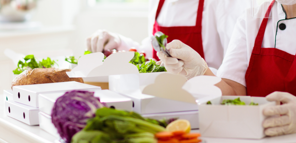Twee medewerkers in een professionele keuken met rode schorten en handschoenen die zorgvuldig verse salades verpakken in witte, kartonnen meeneemdozen.