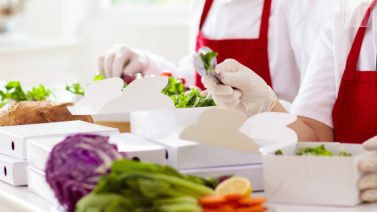 Twee medewerkers in een professionele keuken met rode schorten en handschoenen die zorgvuldig verse salades verpakken in witte, kartonnen meeneemdozen.
