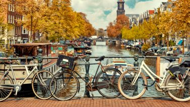 Vier fietsen geparkeerd op een brugleuning langs een gracht in Amsterdam met herfstkleurige bomen en historische gebouwen voor de Werelfietsdag 2025.